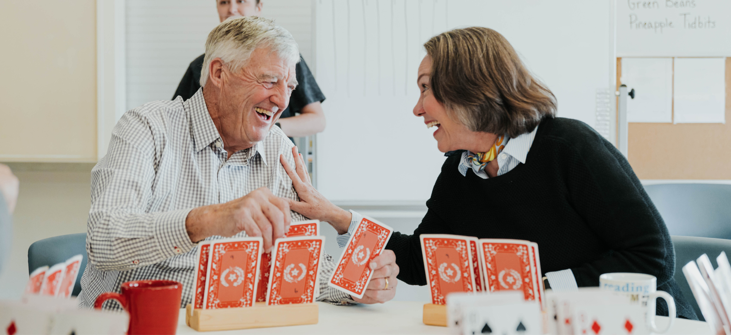 An older man and woman sit at a table playing oversized cards as they laugh together and the woman's hand is on his shoulder.