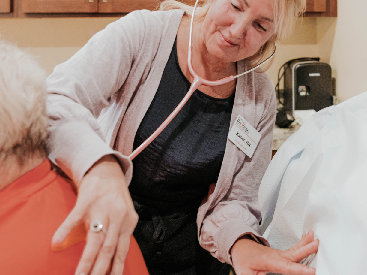 A smiling woman wearing a stethoscope uses it against the back of a person so that she can hear their heartbeat.