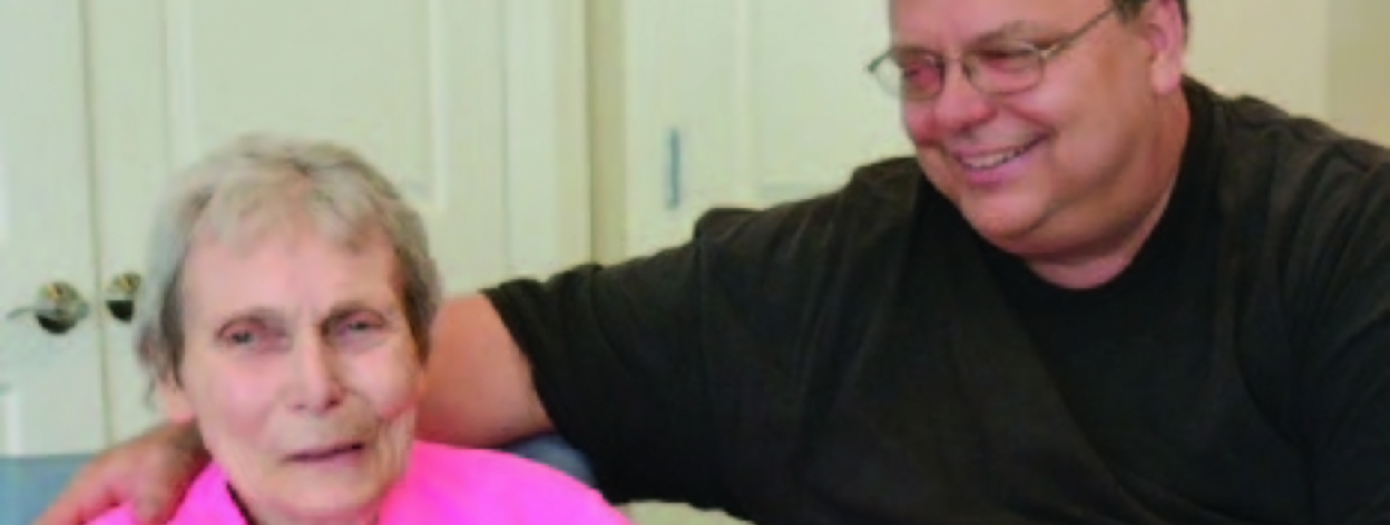 A smiling man wearing glasses has placed his arm around his elderly mother.