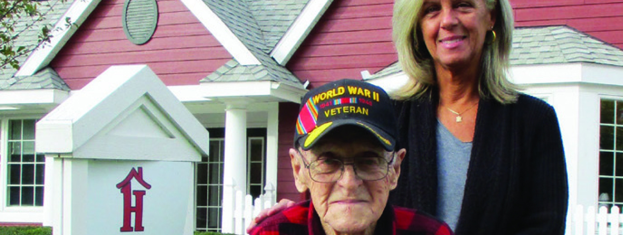 An elderly man with glasses and a baseball cap sits in a wheelchair next to The Little Red House sign as a woman stands behind him.