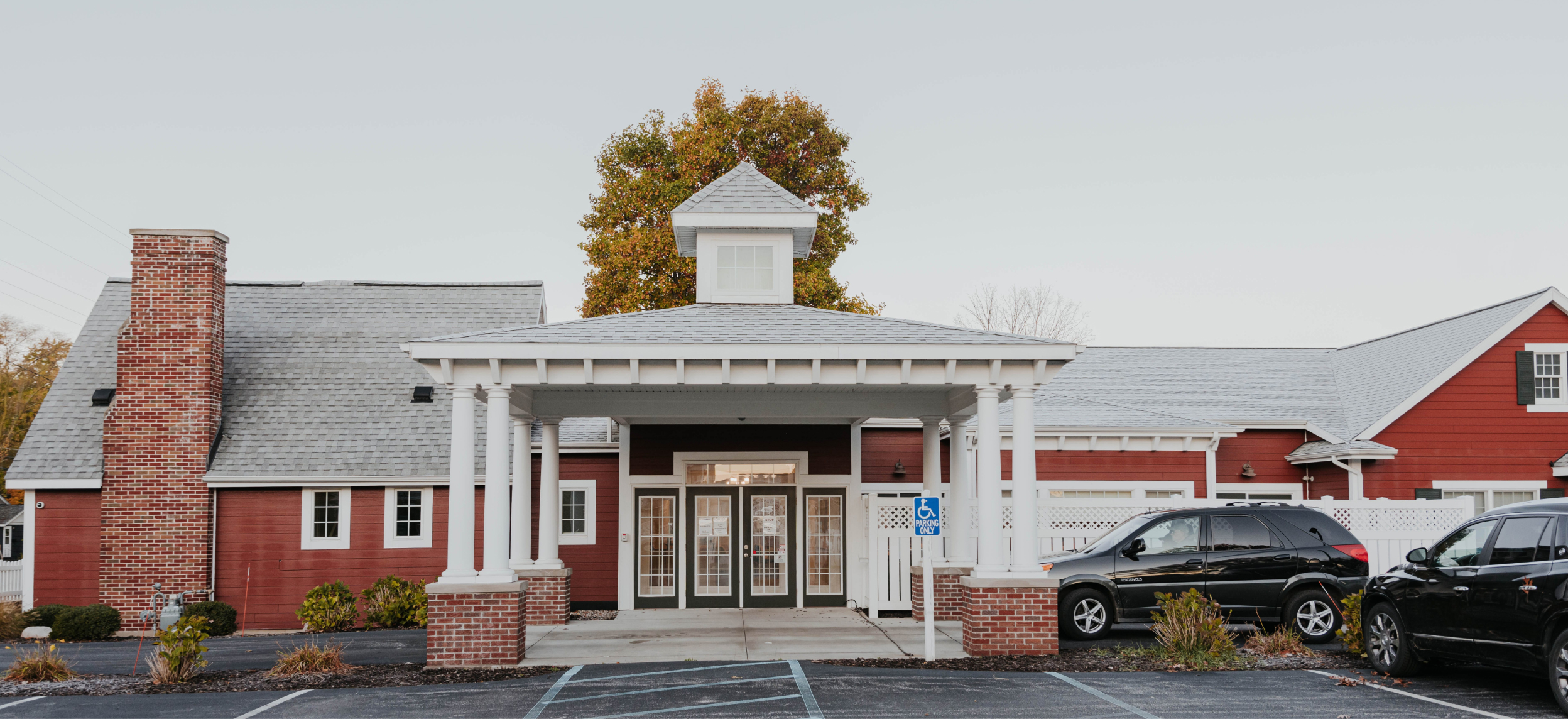 Front entrance and facade of The Little Red House facility showing the columns and a cupola and a car parked in the parking lot.
