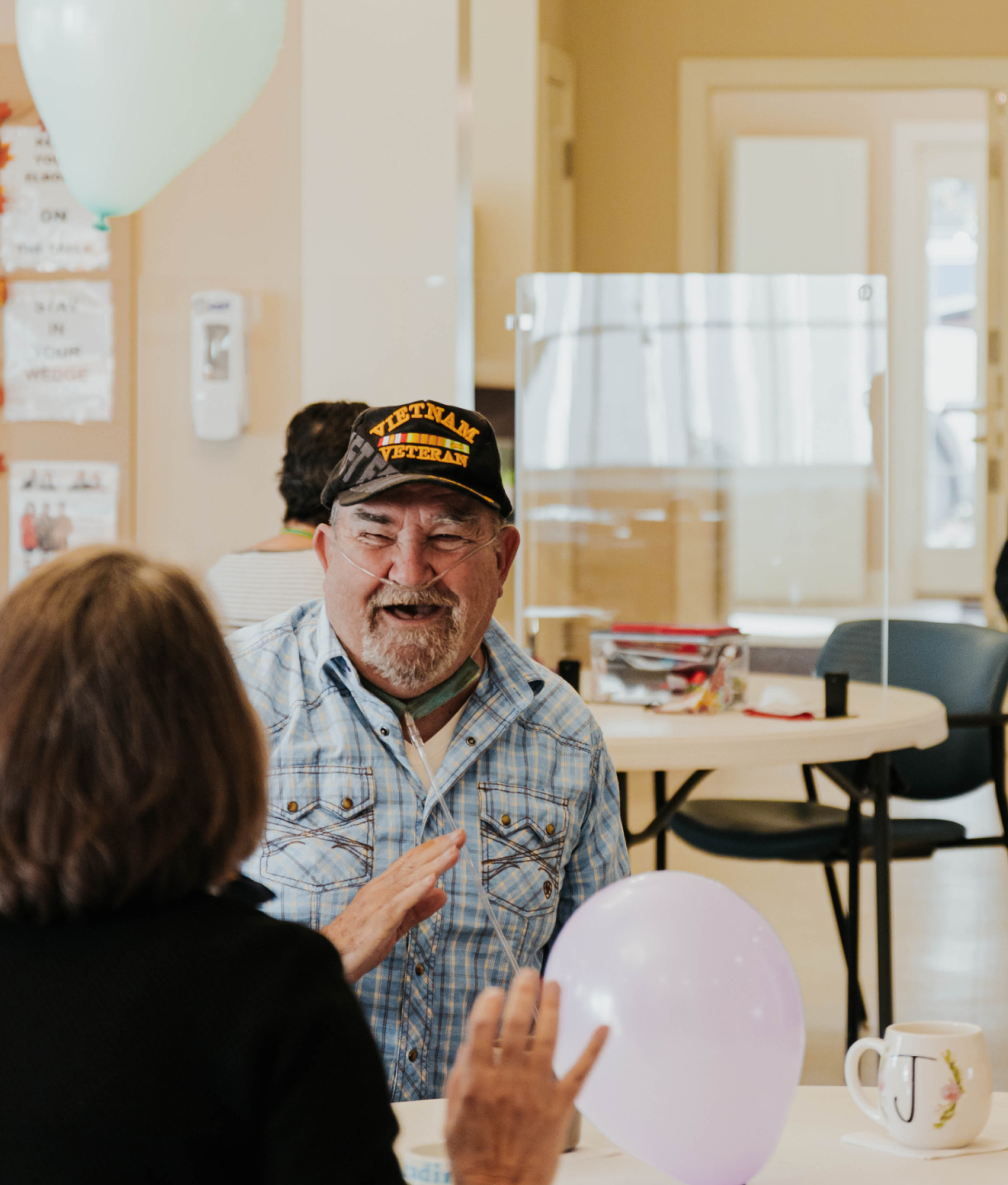 A bearded man with a Vietnam Veteran baseball cap sits at a table facing a woman as a balloon floats above them while another ballon and coffee mug are on the table.