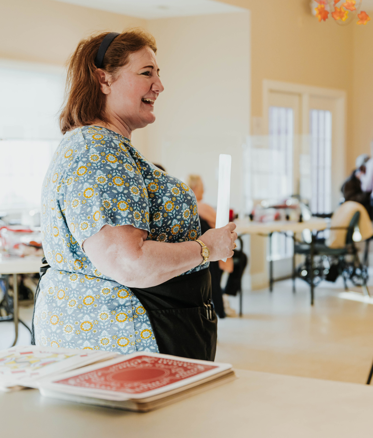 A smiling woman wearing an apron is standing in a room with tables.