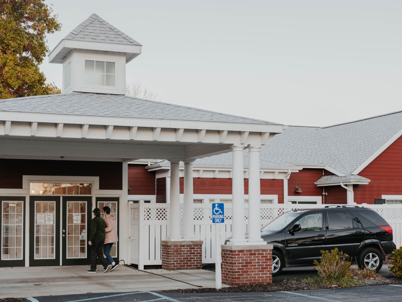 Two people walk into The Little Red House main entrance with a car parked to the side.