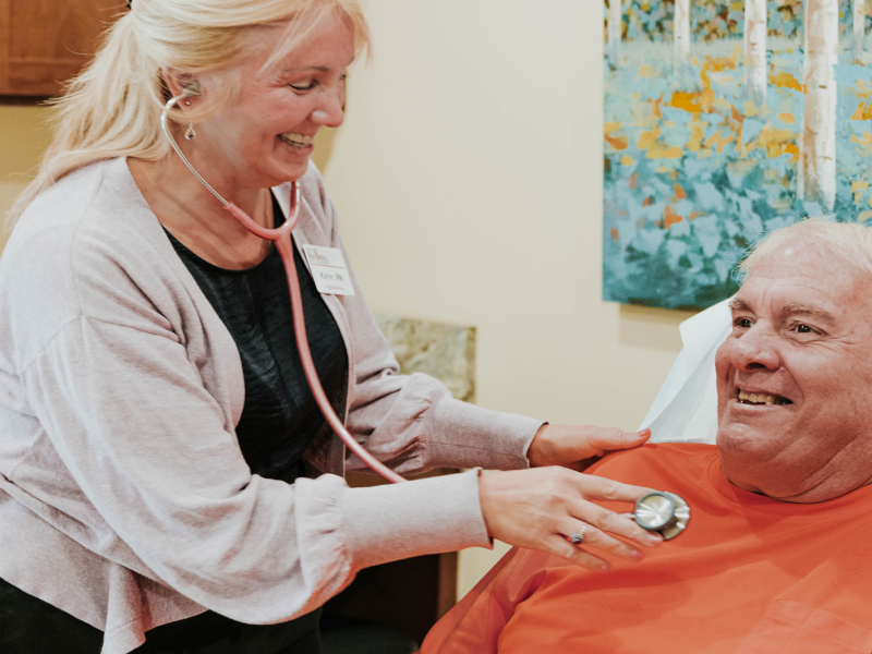 A smiling woman wearing a stethoscope uses it check the heartbeat of a seated and smiling elderly man.