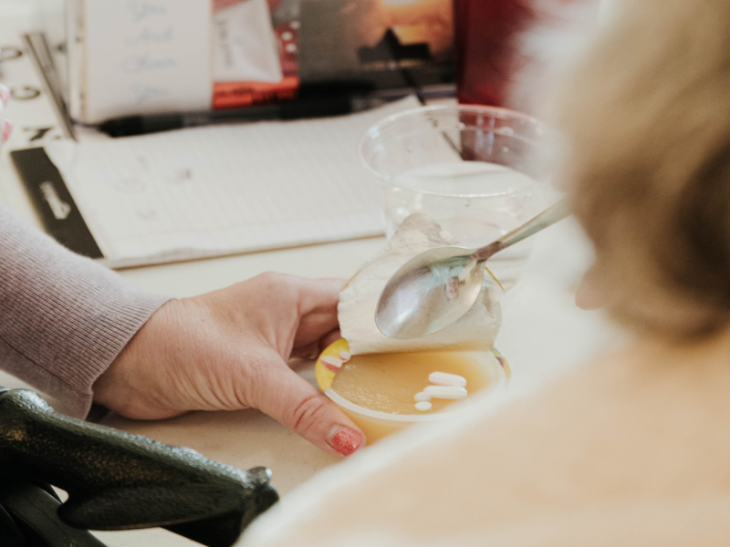 A close-up of a hand holding a cup of applesauce with pills in it as another hand holds a spoon near the container.