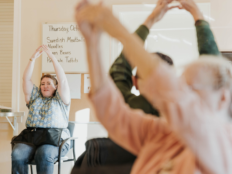 A woman in the background, sitting in front of a sign showing the day's menu of Swedish Meatballs and beans, raises her arms over her head as she instructs exercises for other people who are doing the same movements in the foreground.