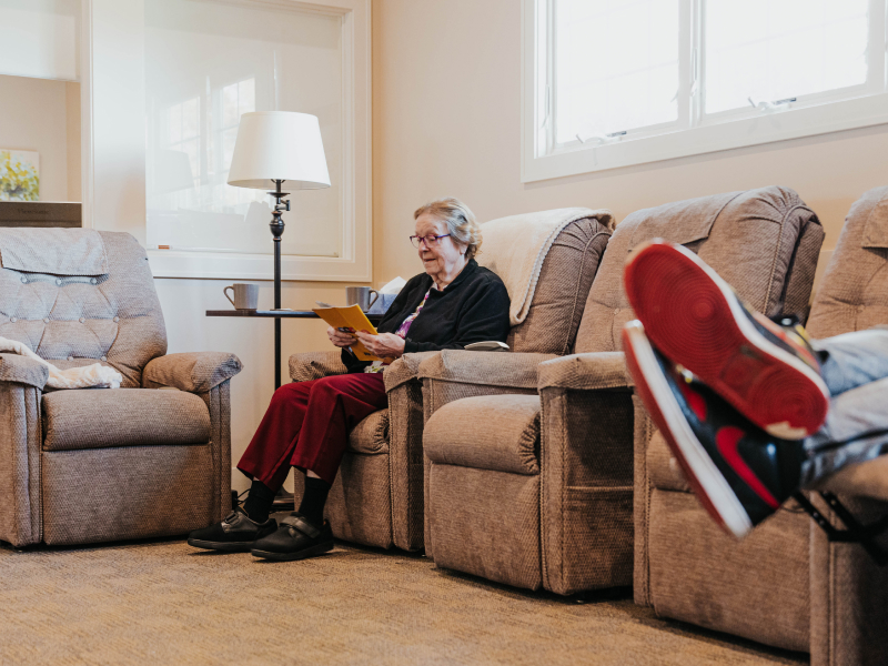 A woman wearing glasses sits on a recliner while reading a book while athletic shoes and crossed legs can also be seen in the foreground.
