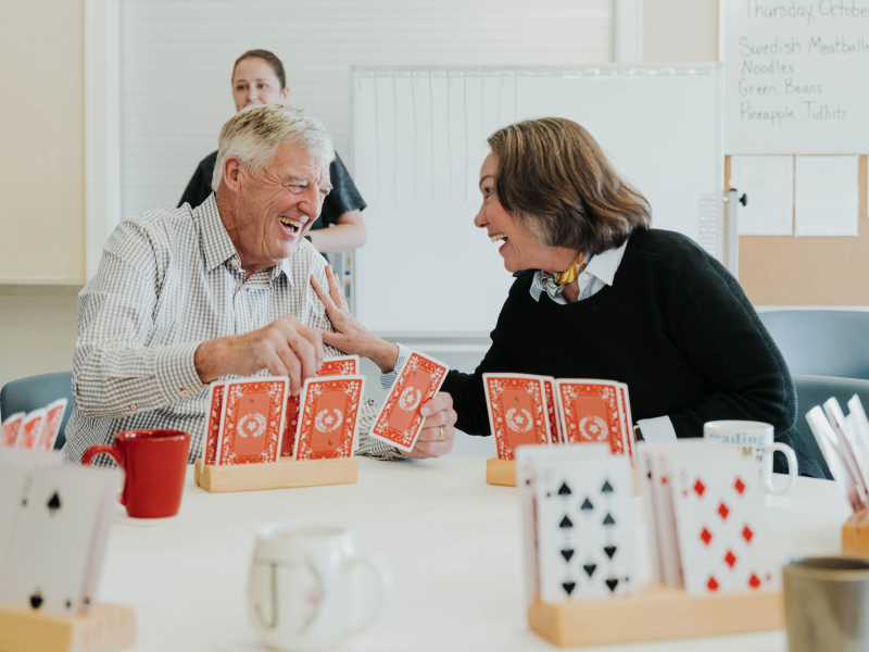 An older man and woman sit at a table playing oversized cards as they laugh together and the woman's hand is on his shoulder.