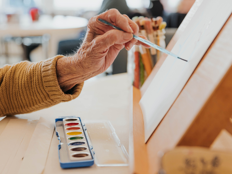 Close-up of an older person's hand holding a paint brush against paper laying against and easel as a box of paints sits below.